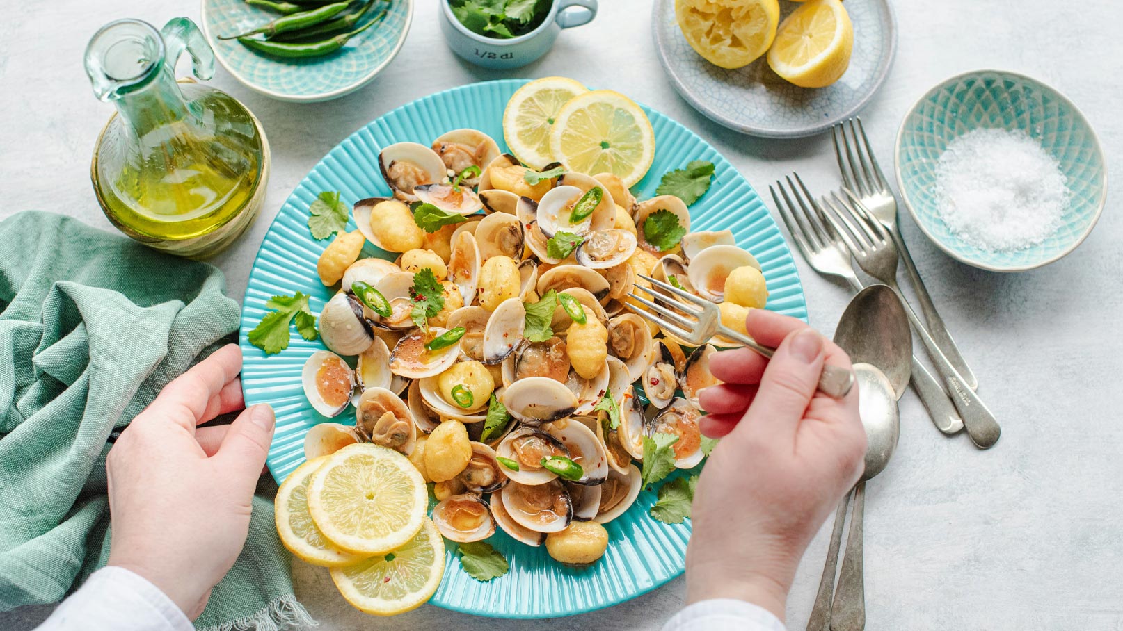 Ein Teller mit Pasta und Muscheln, garniert mit Zitronenscheiben und frischen Kräutern. Eine Hand hält eine Gabel, bereit, das Gericht zu servieren. Daneben sind eine Flasche Olivenöl, eine Schale mit Salz und einen kleinen Behälter mit frischem Koriander sichtbar.