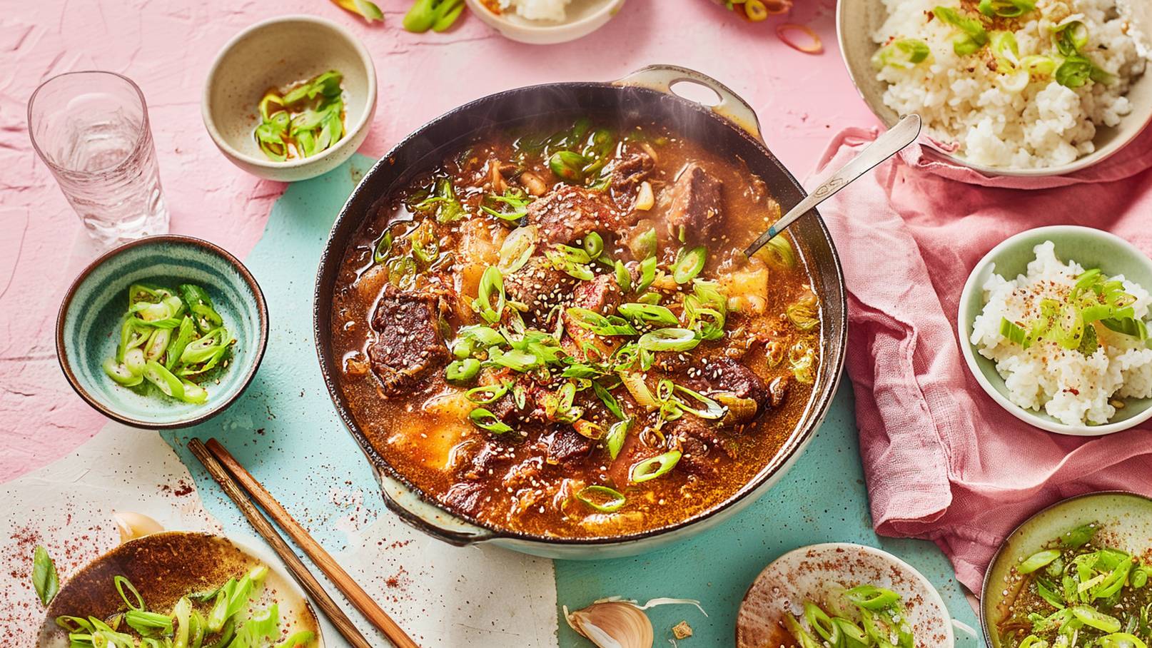 Eine große Schüssel mit dampfendem Nudelgericht, das aus gelben Nudeln, gebratenem Fleisch, Gemüse und einer würzigen Sauce besteht. Reste von Tofustücken liegen neben einer Mischung aus grünem Blattgemüse und Zwiebeln. Das Gericht ist reich an Farben und Texturen.