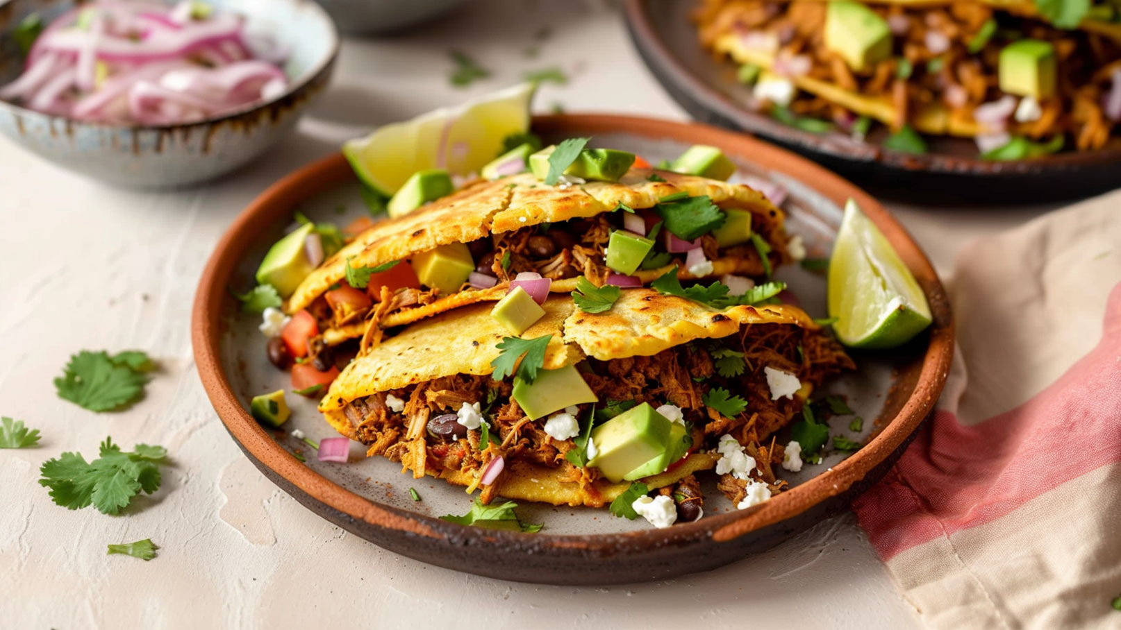 Auf dem Bild sind drei gefüllte Tacos auf einem braunen Teller zu sehen. Die Tacos sind mit zerrissenem Fleisch, frischen Kräutern, Tomaten, roten Zwiebeln und Avocado-Stücken belegt. Eine Limettenscheibe und einige Korianderblätter sind ebenfalls sichtbar.