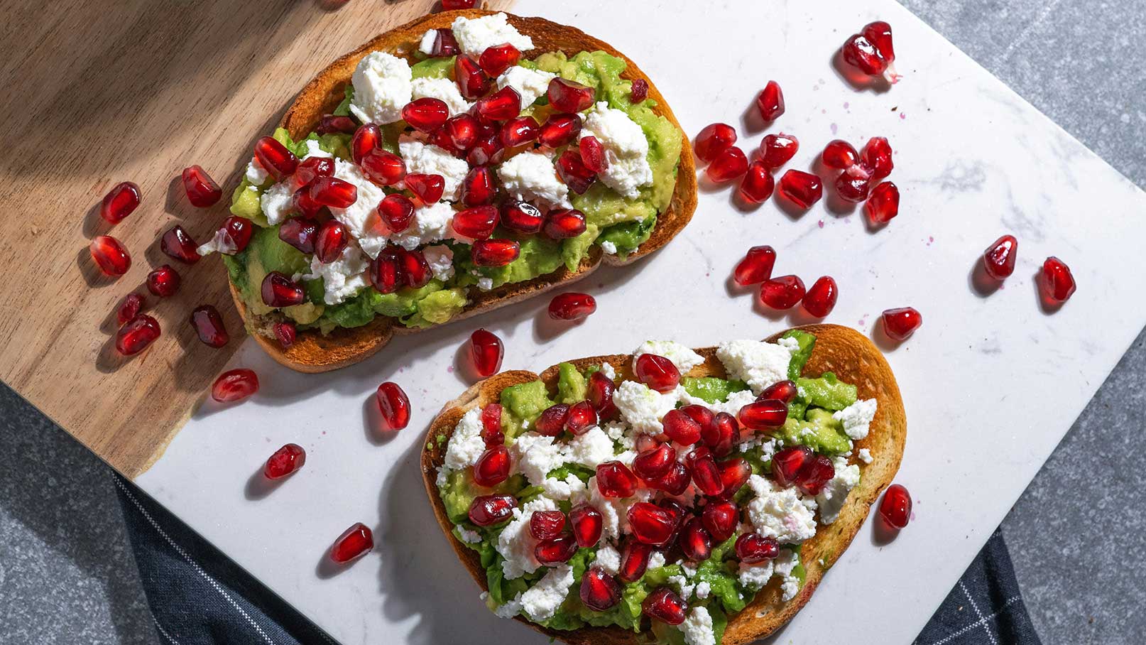 Zwei geröstete Brotscheiben mit einer cremigen Avocado-Aufstrich, garniert mit zerbröckeltem Käse und roten Granatapfelkernen. Der Hintergrund ist eine helle, strukturierte Oberfläche, auf der einige Granatapfelkerne verstreut liegen.