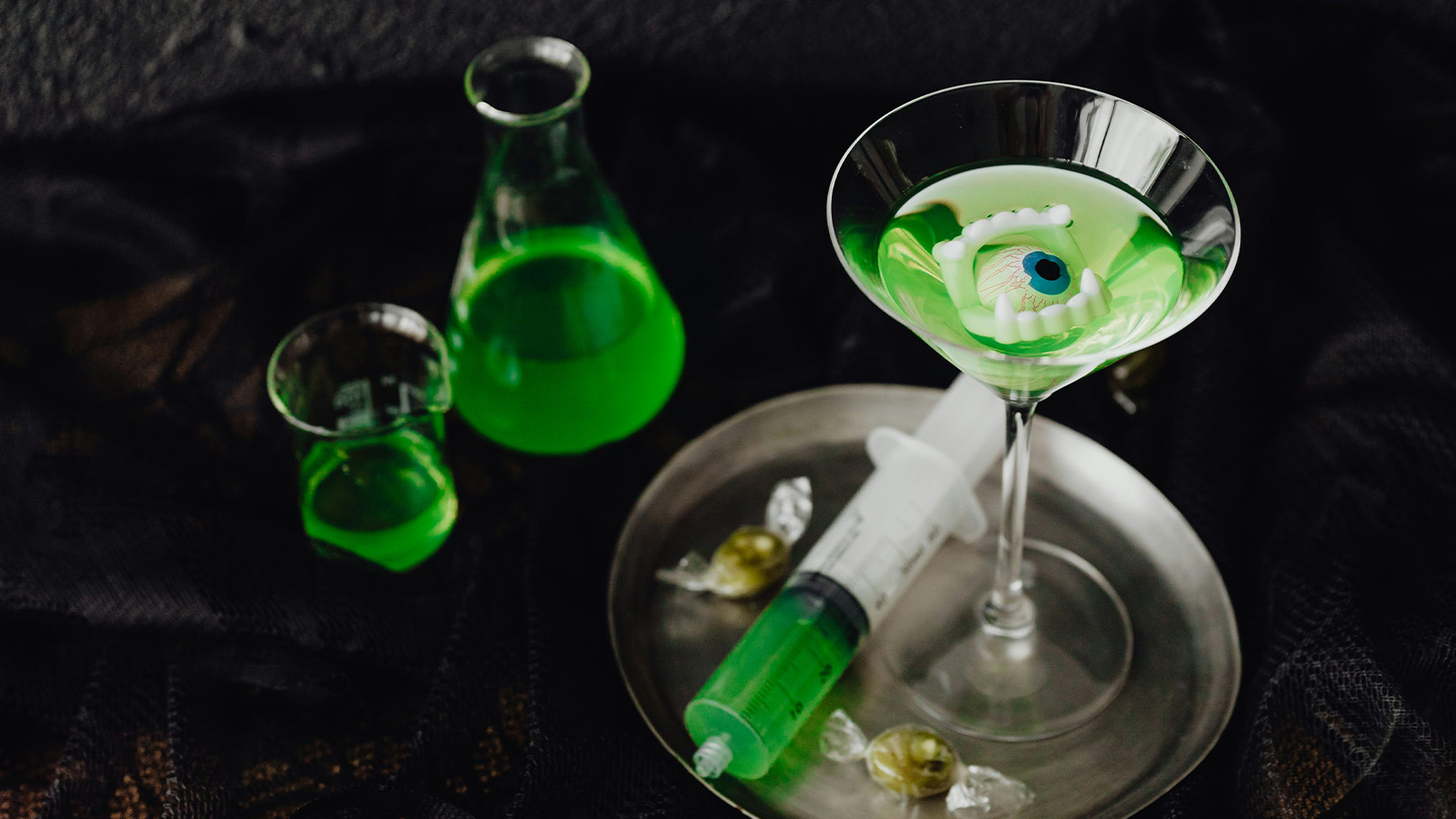 Ein grün gefärbter Cocktail in einem Martini-Glas enthält eine Augenfigur und Zähne. Neben dem Glas stehen verschiedene konische Behälter mit grülicher Flüssigkeit und eine Spritze auf einem silbernen Tablett. Die Szene vermittelt eine unheimliche, experimentelle Atmosphäre.