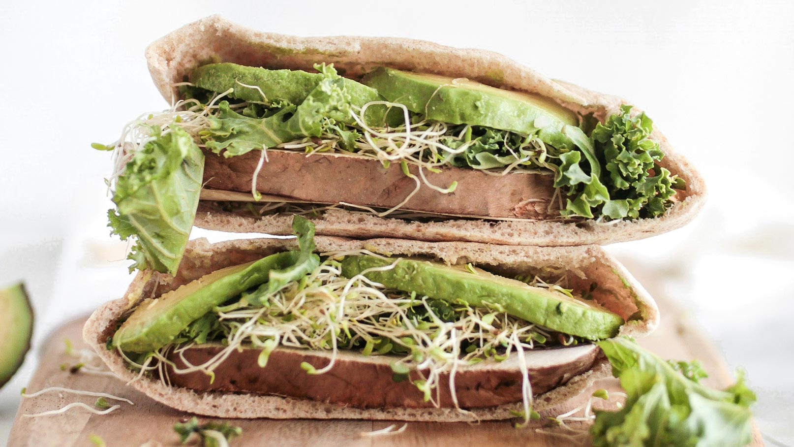 Ein aufgeschnittener Vollkorn-Pita-Brot mit frischem Gemüse. Darin befinden sich Schichten aus Salat, Avocado und Sprossen. Die Füllung ist üppig und grün, was einen gesunden Snack andeutet.