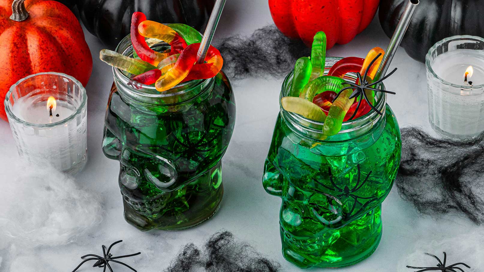 Zwei grüne Getränke in Totenkopf-Gläsern, dekoriert mit Gummibärchen und Spinnen, stehen auf einem Tisch. Im Hintergrund sind Kürbisse und Kerzen platziert. Die Szene hat ein gruseliges Halloween-Thema.