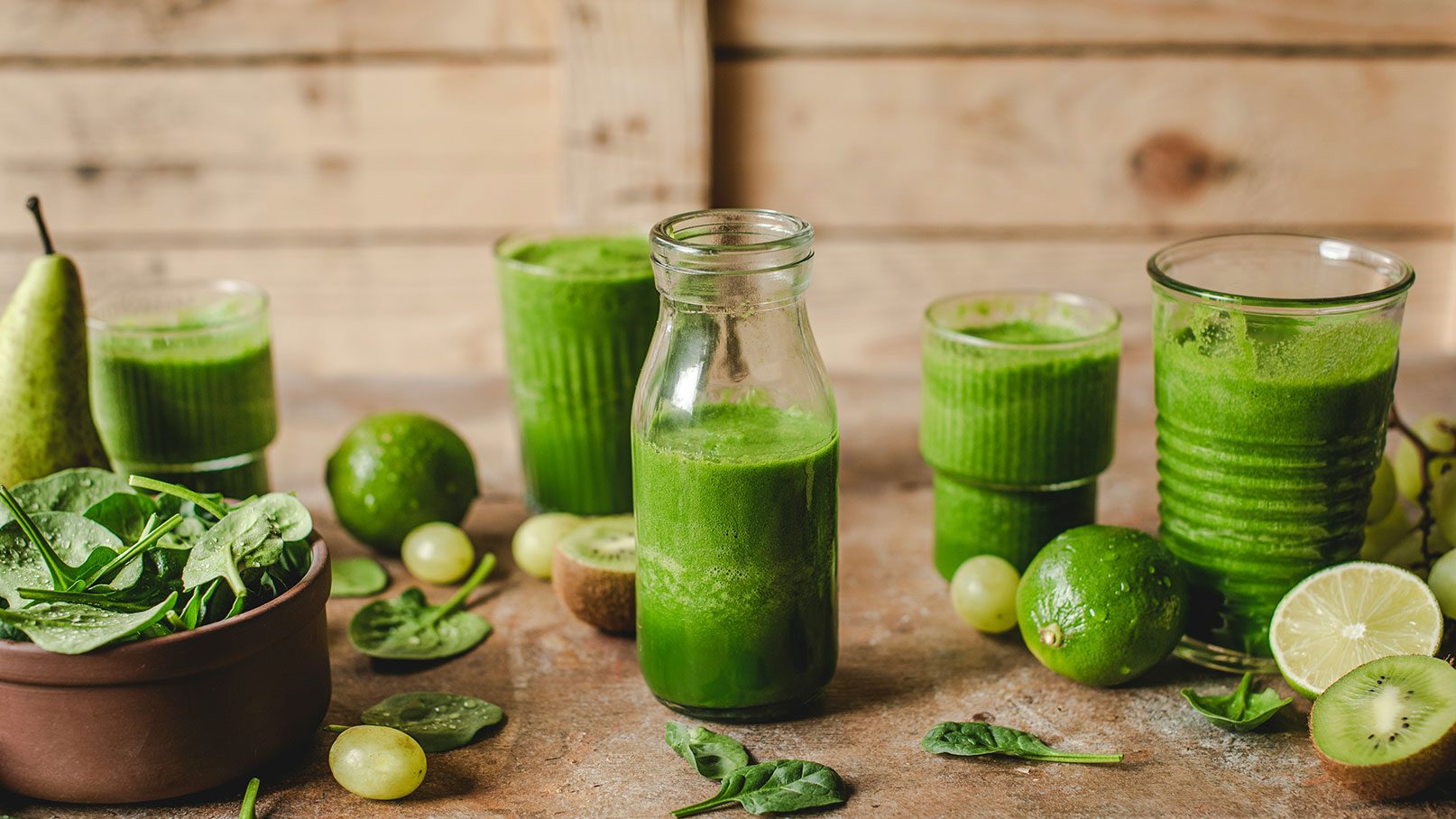 Auf dem Bild sind mehrere grüne Smoothies in Gläsern und einer Flasche angeordnet. Dazu befinden sich frisches Blattspinat, eine Limette, eine Kiwi und eine Birne auf einem Holztisch. Die gesamte Anordnung vermittelt einen gesunden, frischen Eindruck.