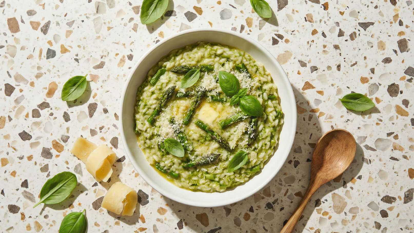 Ein Teller mit grünem Risotto, garniert mit frischem Basilikum und Spargelstücken. Das Risotto hat eine cremige Konsistenz und wird von Parmesanspänen ergänzt. Im Hintergrund ist eine helle Oberfläche aus Terrazzo sichtbar, sowie ein Holzlöffel.
