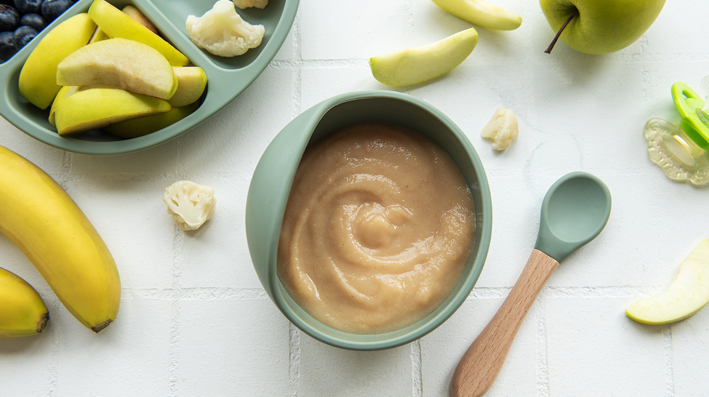 Eine grüne Schüssel mit cremigem Obstbrei steht auf einer weißen Oberfläche. Um die Schüssel sind frische Zutaten wie grüne Äpfel, Bananen und Blumenkohlstücke angeordnet. Ein kleiner Löffel aus Holz und Silikon liegt neben der Schüssel.