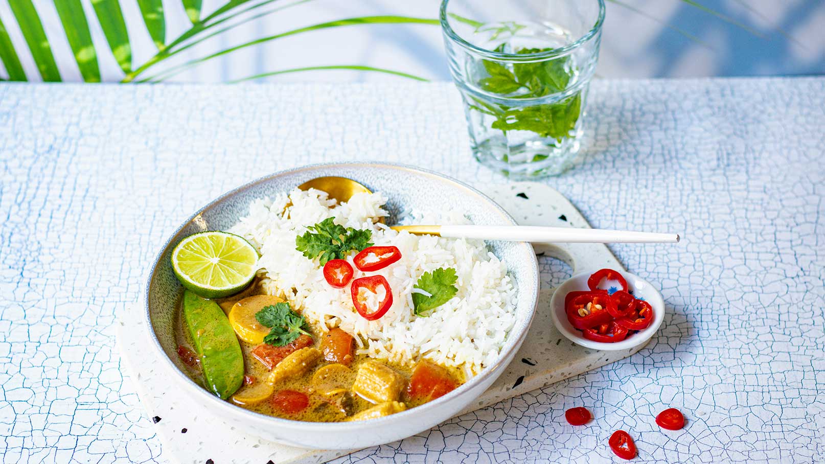 Eine Schale mit Reis und einer bunten Gemüse-Curry-Sauce, garniert mit Koriander, Limettenscheiben und roten Chilischoten. Daneben befindet sich ein Glas Wasser mit Minze. Der Hintergrund zeigt eine pflanzenartige Textur.