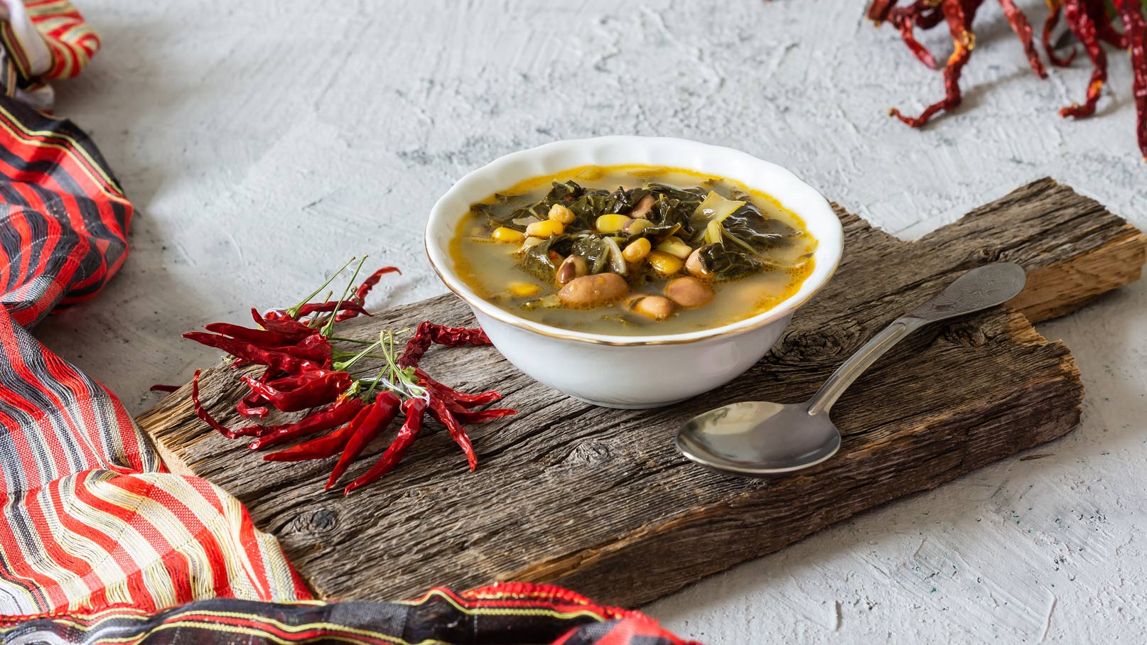 Eine Schüssel mit einer herzhaften Suppe auf einem Holzbrett. Die Suppe enthält Bohnen, Mais und Grünkohl. Rote getrocknete Chilischoten liegen neben der Schüssel. Ein Löffel aus Metall ist ebenfalls sichtbar.