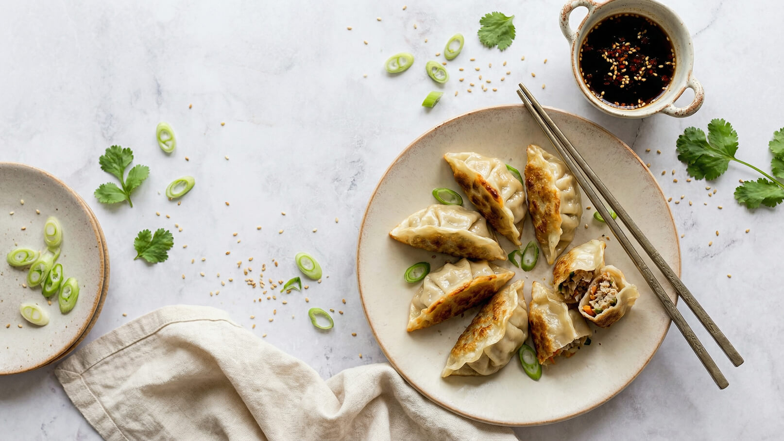Ein Teller mit gebratenen Teigtaschen, umgeben von Frühlingszwiebeln und frischem Koriander. Eingerichtet sind ebenfalls Essstäbchen und eine Schüssel mit dunkler Soße. Die Teigtaschen sind goldbraun und zeigen ihre Füllung.