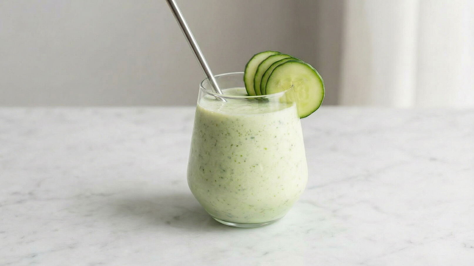 Ein Glas mit einem erfrischenden, grünen Smoothie steht auf einem hellen Tisch. Der Smoothie hat eine cremige Konsistenz und ist mit einer Scheibe Gurke dekoriert. Ein Strohhalm aus Edelstahl steckt im Glas.