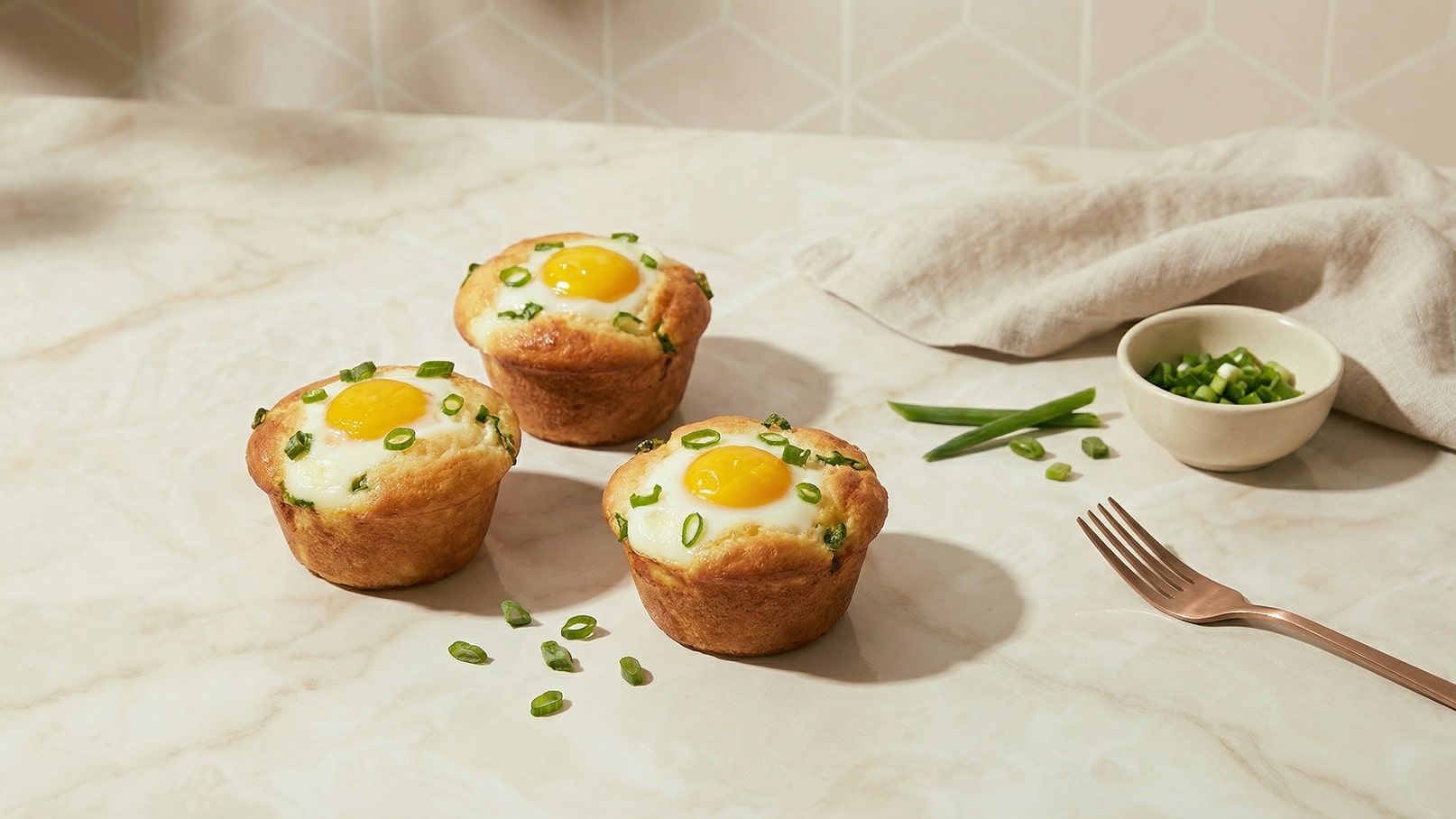 Drei gebackene Muffins sind auf einer hellen Oberfläche angerichtet. Jeder Muffin ist mit einem Ei und frischen Schnittlauchstücken befüllt. Neben den Muffins liegt eine kleine Schüssel mit gehacktem Schnittlauch und ein Essbesteck.