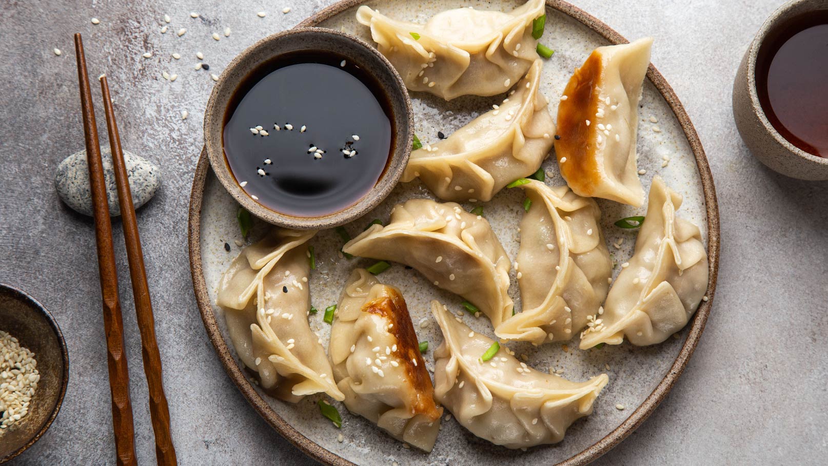 Ein Teller mit dampfend heißen Dumplings, garniert mit Sesam und Frühlingszwiebeln. Daneben befindet sich eine kleine Schale mit dunkler Sojasauce. Zwei Essstäbchen liegen auf der Seite des Tellers. Die Dumplings sind goldbraun und haben eine appetitliche Form.