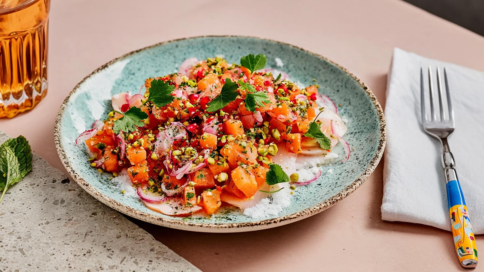 Ein Teller mit frischem Salat aus gewürfelten Melonen, Zwiebeln und gehackten Pistazien. Der Salat ist mit frischen Kräutern garniert und hat ein lebhaftes, farbenfrohes Aussehen. Daneben liegt ein Besteckset und ein Glas mit einem Getränk.