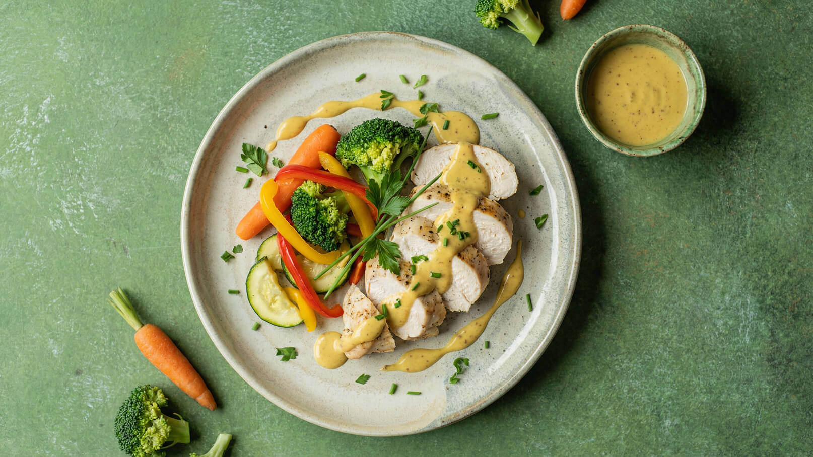 Ein Teller mit zartem, geschnittenem Hähnchenbrustfilet in einer cremigen Soße, garniert mit frischer Petersilie. Neben dem Fleisch liegen bunte Gemüsestreifen aus Paprika, Brokkoli, Karotten und Zucchini. Ein kleiner Schälchen mit Soße ist ebenfalls vorhanden.
