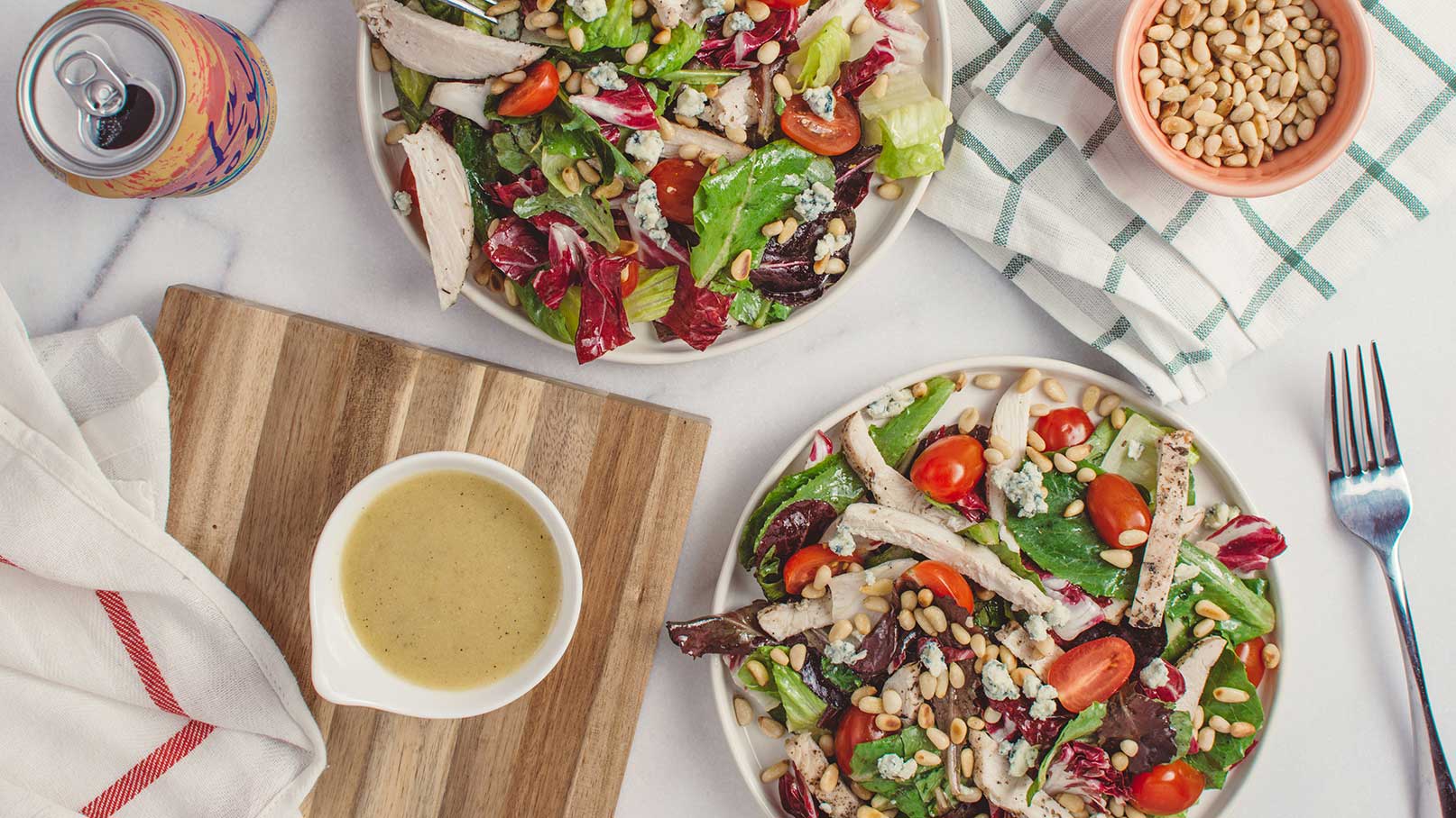 Zwei Teller mit buntem Salat sind auf einem Tisch arrangiert, einer mit Hähnchenstücken, Kirschtomaten, Pinienkernen und Blauschimmelkäse. Daneben steht eine kleine Schüssel mit Salatsoße. Ein Holzbretter und eine Gabel sind ebenfalls sichtbar.