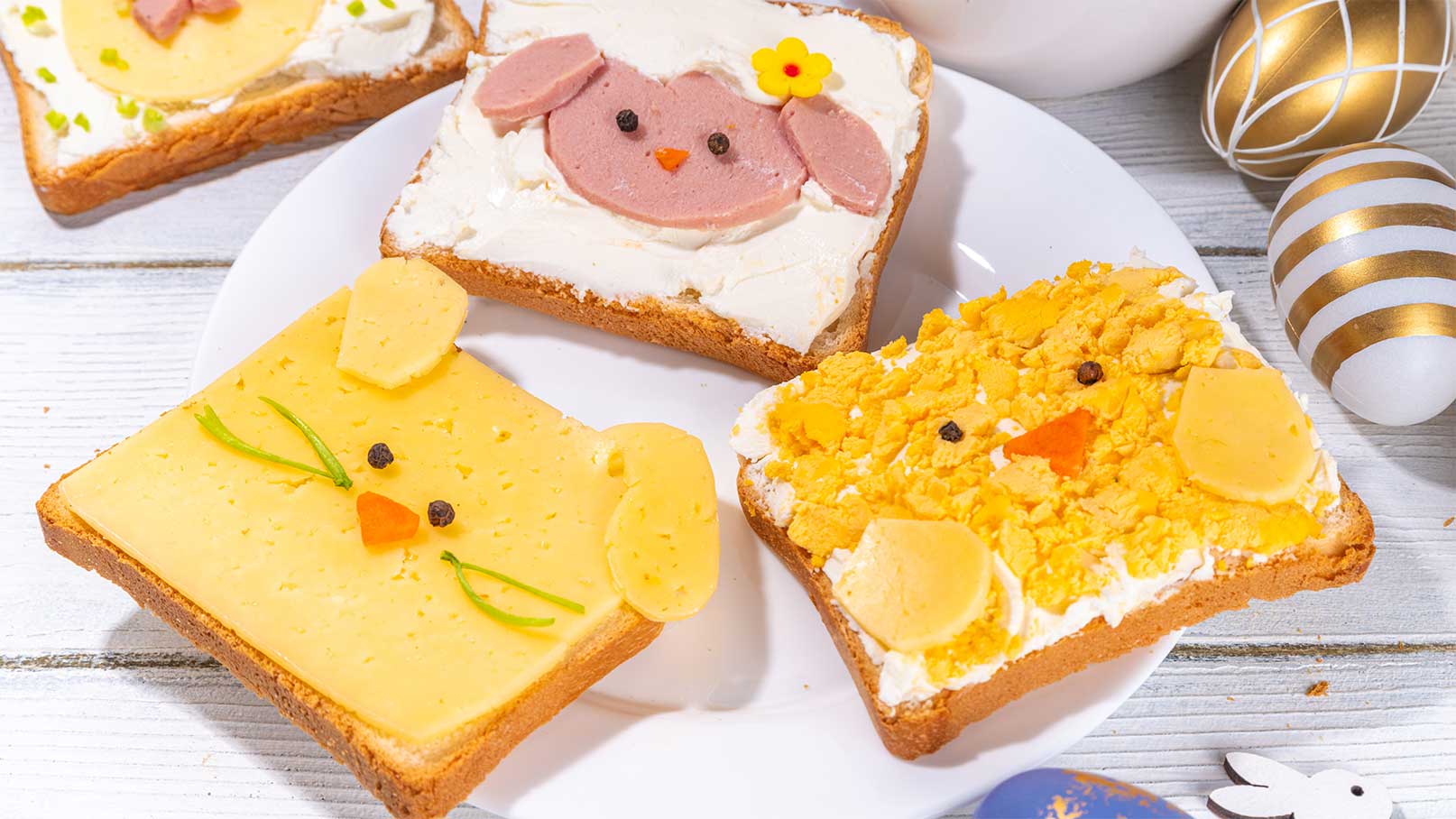Auf einem weißen Teller liegen mehrere belegte Brote, die speziell dekoriert sind. Jedes Brot hat ein tierisches Gesicht: ein gelbes Küken, ein braunes Lamm mit Blüte und ein orangefarbenes Huhn. Die Brote sind bunt und ansprechend für festliche Anlässe gestaltet.