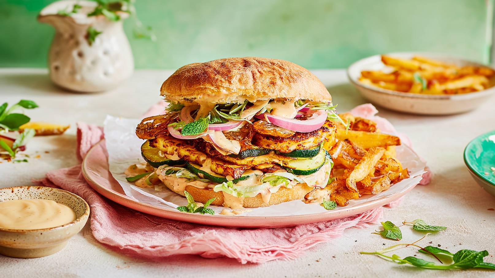 Ein üppiger Burger aus gegrilltem Gemüse, darunter Zucchini und Tortilla, geschichtet mit frischen Kräutern und Zwiebeln. Der Burger liegt auf einem rosa Teller, begleitet von knusprigen Pommes und einer kleinen Schale mit Sauce. Der Hintergrund ist in sanften Grüntönen gehalten.