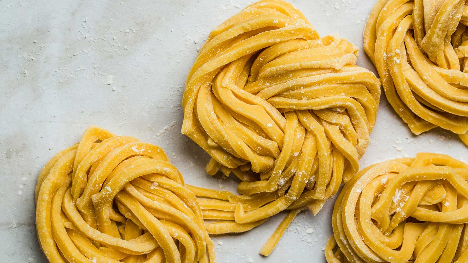 Auf dem Bild sind mehrere Portionen frischer, ungekochter Pastateig abgebildet. Die langen, gelben Teigstreifen sind kunstvoll zu Nestern gedreht und leicht mit Mehl bestäubt. Der Hintergrund ist neutral, um die Pasta hervorzuheben.