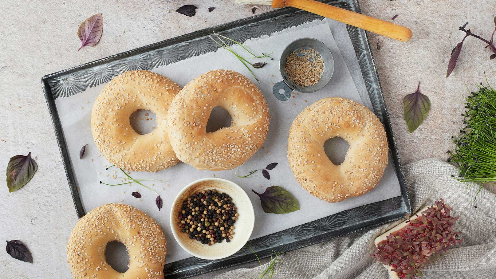 Ein frisch zubereiteter Bagel auf einem Holzbrett, belegt mit knackigem Salat, Tomaten, Gurken und Zwiebeln. Der Bagel hat eine Sesamkruste und ist auf einem Teller angerichtet. Im Hintergrund sind weitere ungeschnittene Bagels sichtbar.