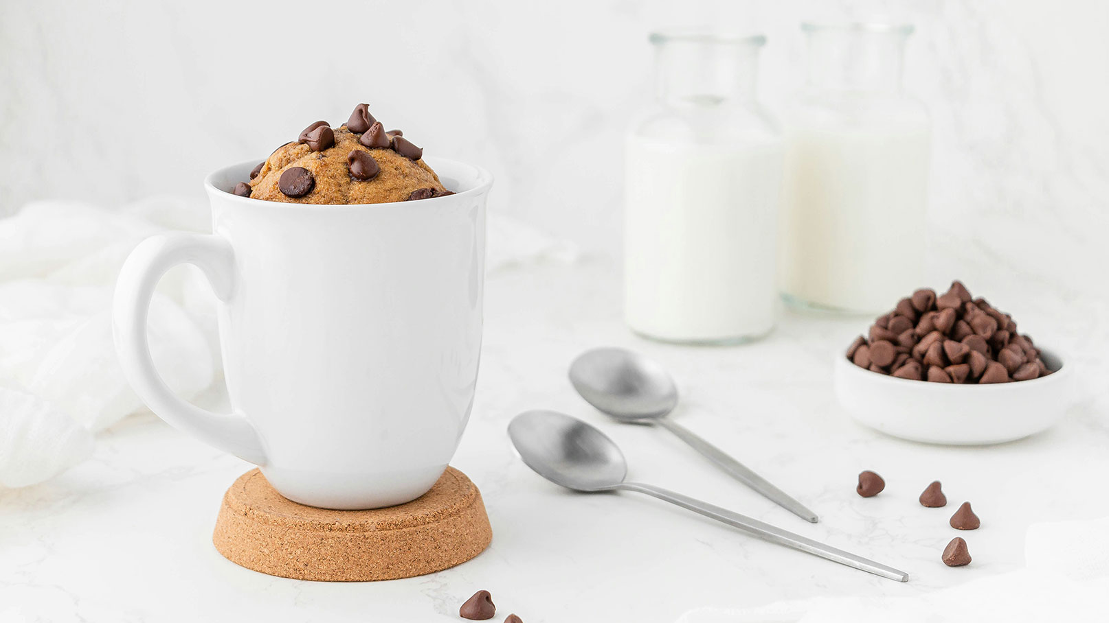 Eine weiße Tasse auf einem Untersetzer enthält einen frisch gebackenen Keks, der mit Schokoladenstückchen belegt ist. Im Hintergrund stehen zwei Flaschen Milch und eine Schüssel mit weiteren Schokoladenstückchen. Zwei Löffel liegen neben der Tasse.