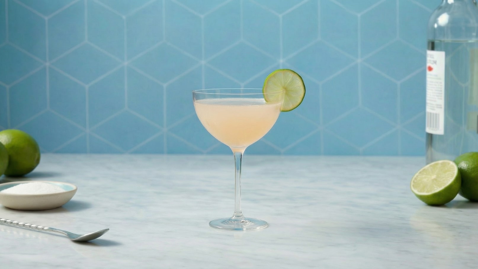 Ein Cocktail in einem eleganten Glas, gefüllt mit einem hellen, leicht trüben Getränk. Eine Limettenscheibe ist am Rand des Glases platziert. Im Hintergrund sind grüne Limetten und ein kleiner Teller mit Zucker zu sehen, vor einer hellblauen, strukturierten Wand.