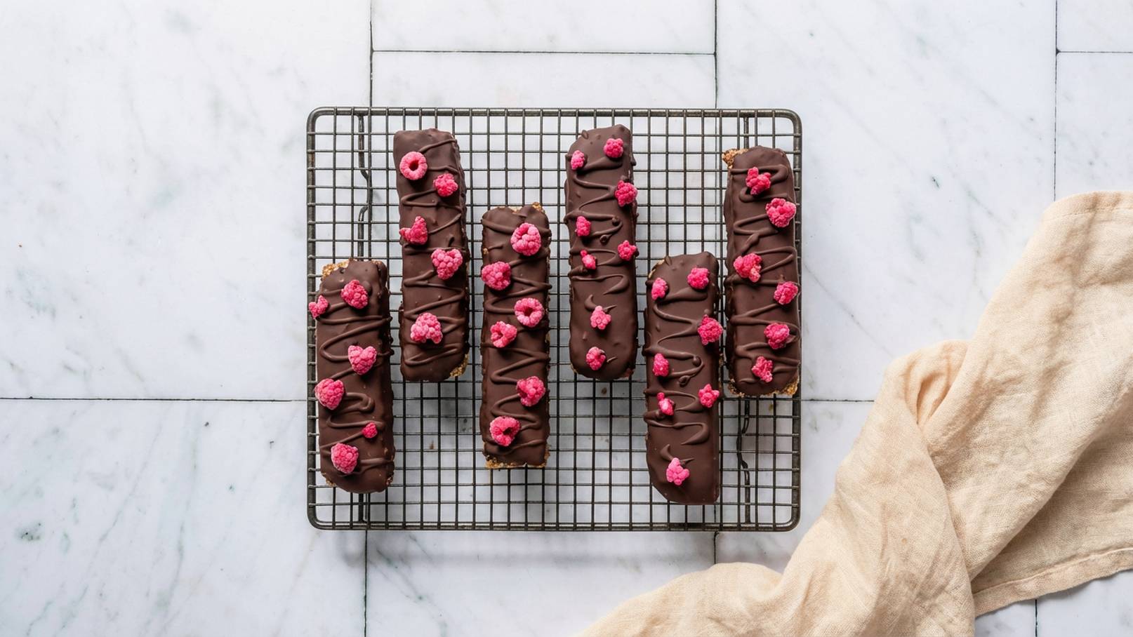 Auf einem Gestell aus Metall liegen mehrere Schokoladenriegel, die mit pinken Himbeeren und Schokoladenguss dekoriert sind. Der Hintergrund zeigt einen hellen, marmorierten Tisch.