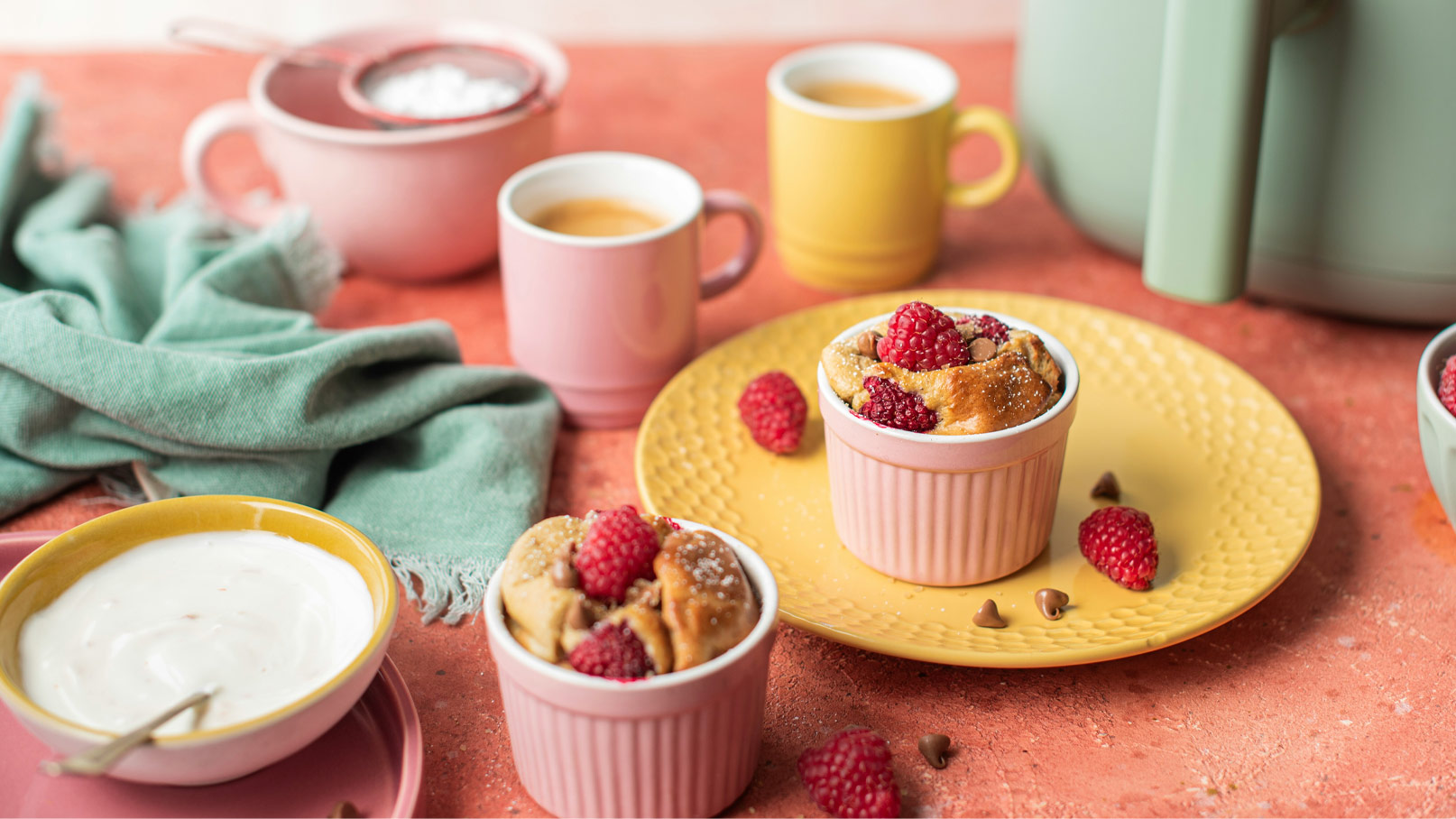Auf dem Bild sind zwei kleine Auflaufformen mit Gebäck und frischen Himbeeren zu sehen. Daneben steht eine große Gelbe Untertasse, auf der sich eine der Formen befindet. Im Hintergrund sind Tassen und ein Tuch sichtbar. Das Gesamtbild vermittelt eine einladende Frühstücksszene.