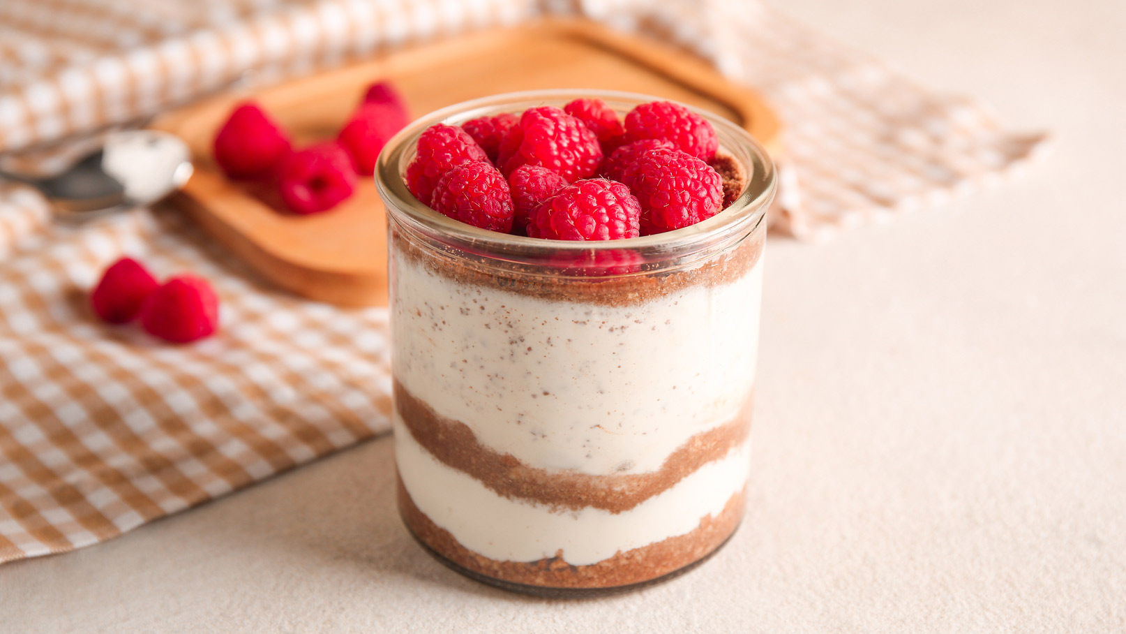 Ein Glas Dessert mit mehreren Schichten: die unterste Schicht besteht aus Schokolade, gefolgt von einer cremigen Vanilleschicht, und oben ist es mit frischen Himbeeren garniert. Im Hintergrund sind einige lose Himbeeren und ein Holzbrett zu sehen.