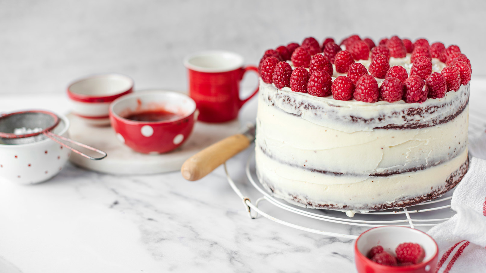Eine zweistöckige Torte mit einer glatten weißen Buttercreme und frischen Himbeeren oben drauf. Rundherum sind einige rote Schalen und einen kleinen Sieb mit Zucker. Der Hintergrund ist unscharf und konzentriert sich auf die Torte im Vordergrund.