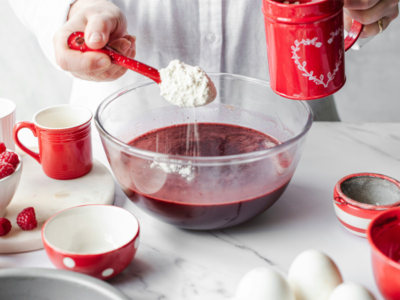 Eine Person hält einen roten Löffel mit Mehl über eine große Schüssel mit roter Flüssigkeit. Um die Schüssel sind verschiedene Küchenutensilien und Zutaten wie Tassen, Schalen und frische Himbeeren angeordnet.