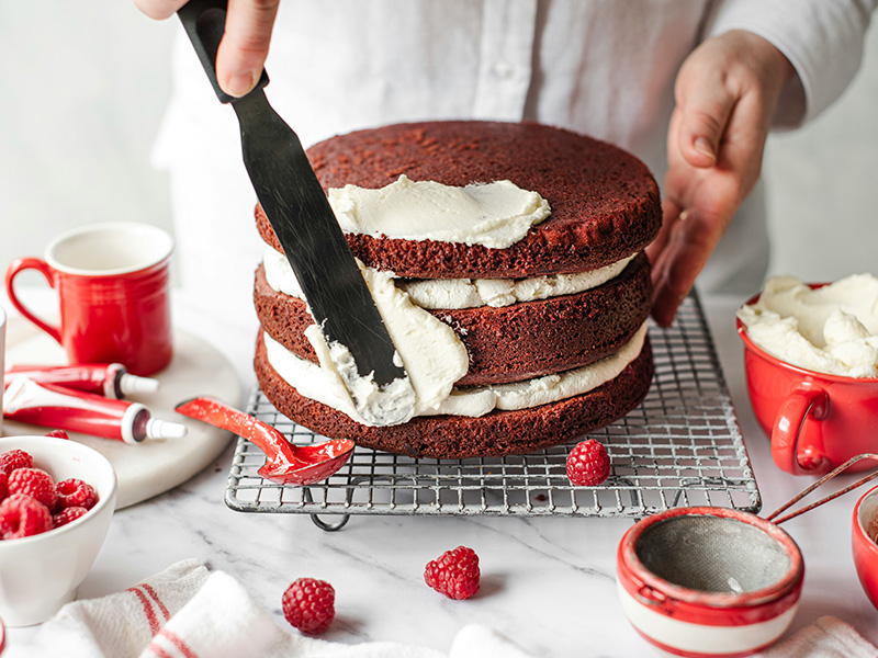 Eine Person schichtet einen roten Samtkuchen mit einer cremigen Füllung. Mit einem Spatel wird die Creme gleichmäßig zwischen den drei Kuchenschichten verteilt. Rote Himbeeren und bunte Dekorationstube liegen auf einem Tisch in der Nähe.