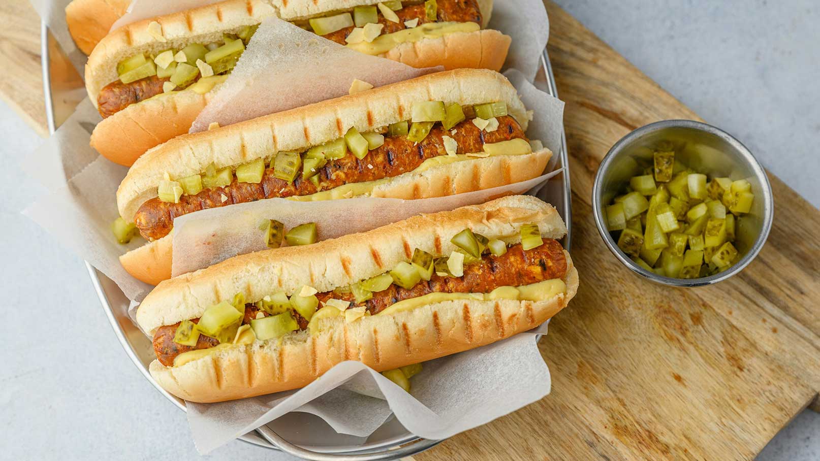 Ein Teller mit mehreren Hotdogs, gefüllt mit einer Wurst und belegt mit gehacktem Gurken. Die Hotdogs sind in Papier gewickelt und liegen in einem metallenen Behälter. Neben dem Teller befindet sich eine kleine Schale mit weiteren gehackten Gurkenstücken.