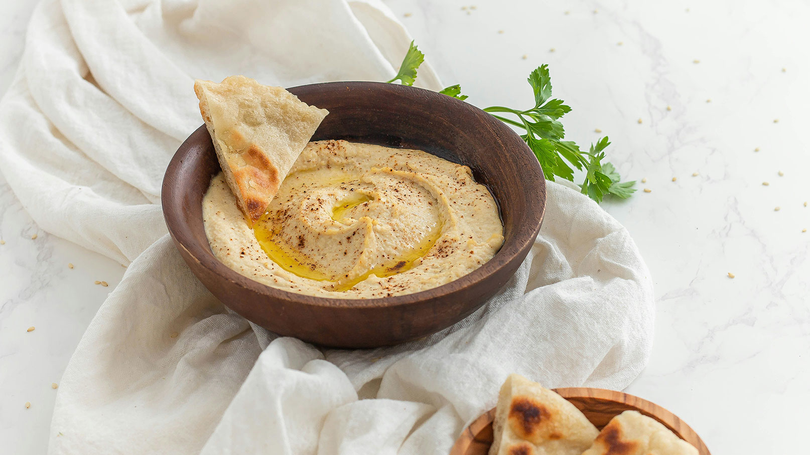 Eine Schüssel mit cremigem Hummus, garniert mit Olivenöl und gewürzten Mustern, steht auf einem hellen Tisch. Neben der Schüssel befindet sich ein Stück Fladenbrot, das in den Hummus getunkt wird. Frische Kräuter sind ebenfalls sichtbar.
