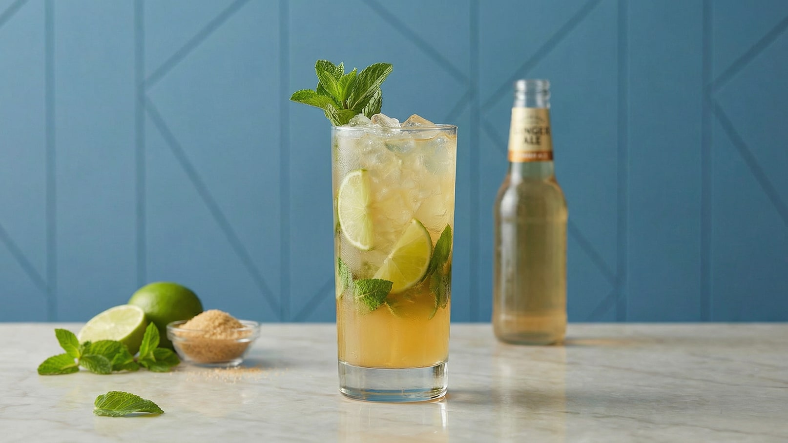 Ein erfrischendes Getränk mit Limettenscheiben und frischer Minze in einem hohen Glas mit Eis. Im Hintergrund steht eine Flasche Ginger Ale. Daneben liegen eine halbe Limette und ein Schälchen mit braunem Zucker. Der Hintergrund ist in einem sanften Blau gehalten.