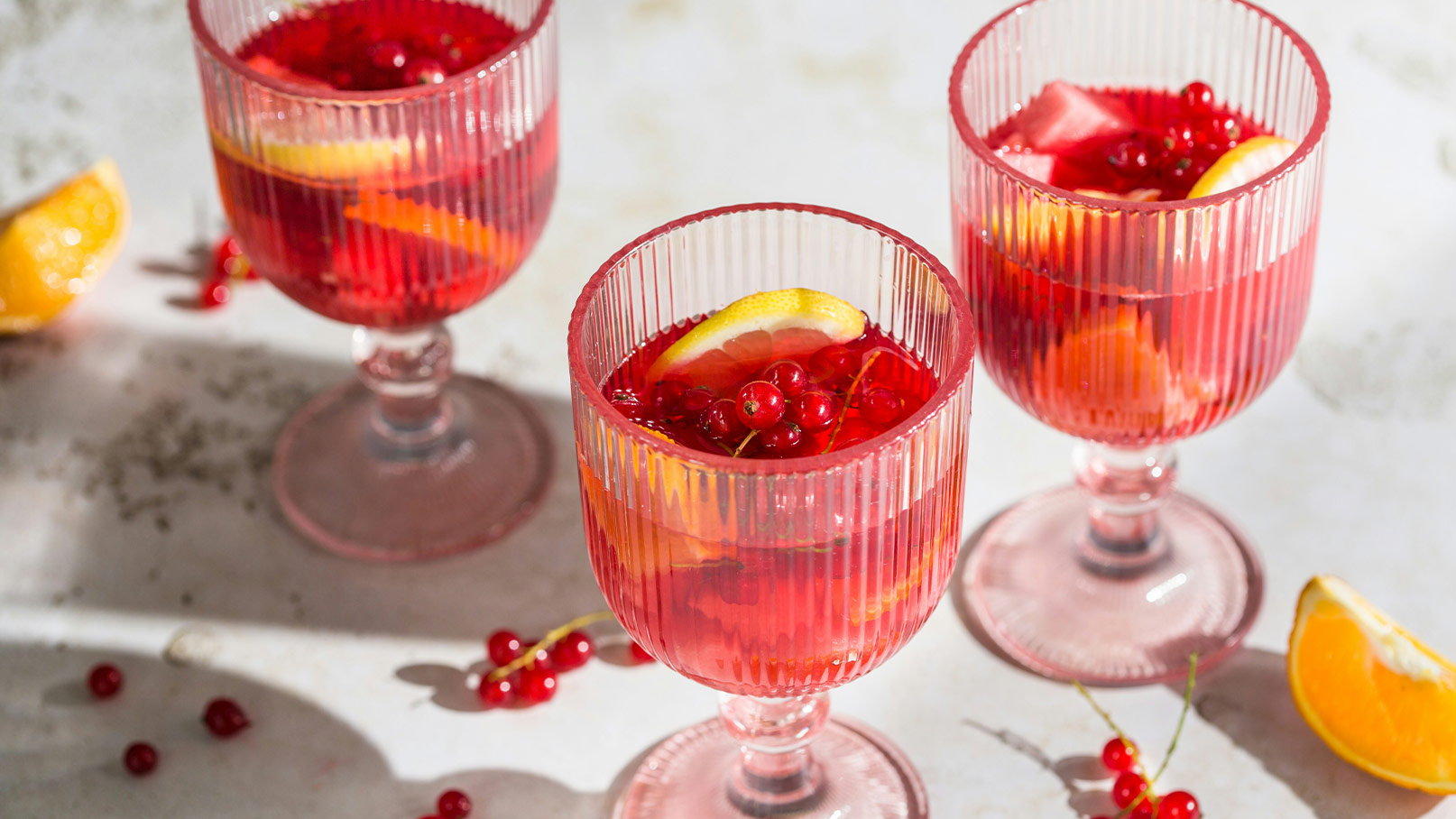 Drei Getränke in gestreiften Gläsern stehen auf einem Tisch. Die Cocktails sind rot und enthalten Stücke von Zitrusfrüchten sowie rote Johannisbeeren. Neben den Gläsern liegen einige frische, orangefarbene Stücke Zitrone und rote Beeren.
