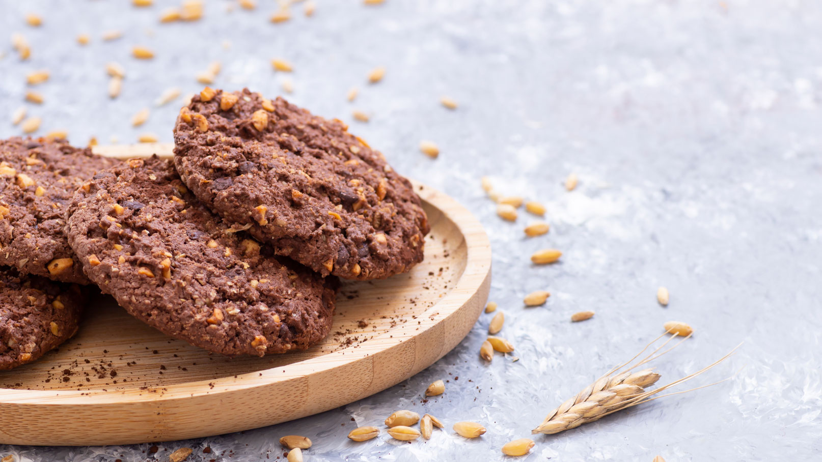 Ein Holzbrett mit mehreren schokoladigen Keksen, die mit Nüssen und Haferflocken verfeinert sind. Im Hintergrund liegen einige Haferkörner verstreut. Die Kekse sind knusprig und haben eine rustikale, appetitliche Erscheinung.