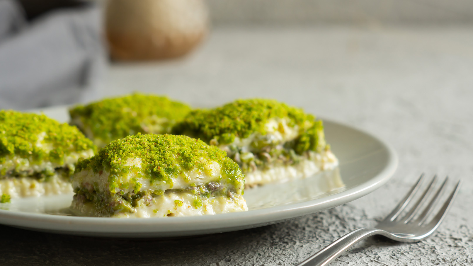 Ein Teller mit mehreren Schichten Dessert, das mit grünem Pistazienpulver bedeckt ist. Die Schichten bestehen aus einer cremigen Füllung und einer knusprigen Basis. Eine Gabel liegt neben dem Teller.