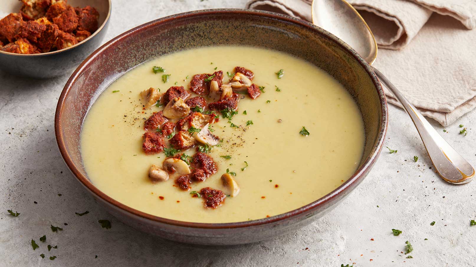 Eine cremige Suppe in einer Schüssel, garniert mit gehackten Nüssen und frischen Kräutern. Ein Löffel liegt daneben. Im Hintergrund ist eine kleine Schüssel mit einer knusprigen Beilage zu sehen.