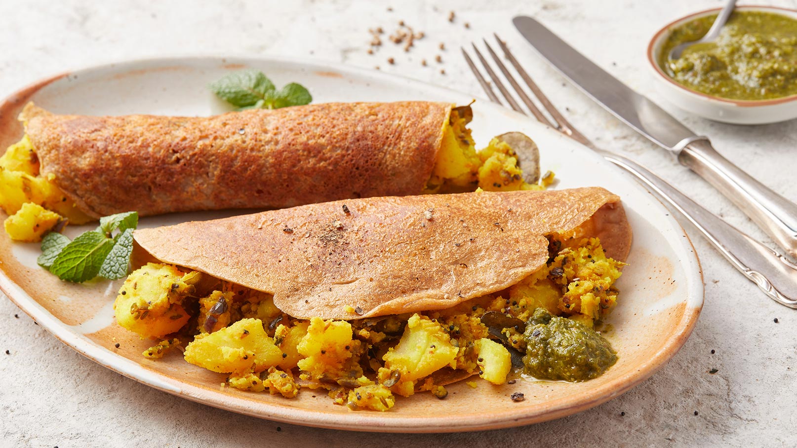 Auf dem Bild sind zwei gefüllte Dosas zu sehen, die auf einem Teller serviert werden. Die Dosas sind aus braunem Teig und sind mit gewürzten Kartoffeln gefüllt. Neben den Dosas liegt etwas grüne Soße. Frische Minzblätter dekorieren den Teller.