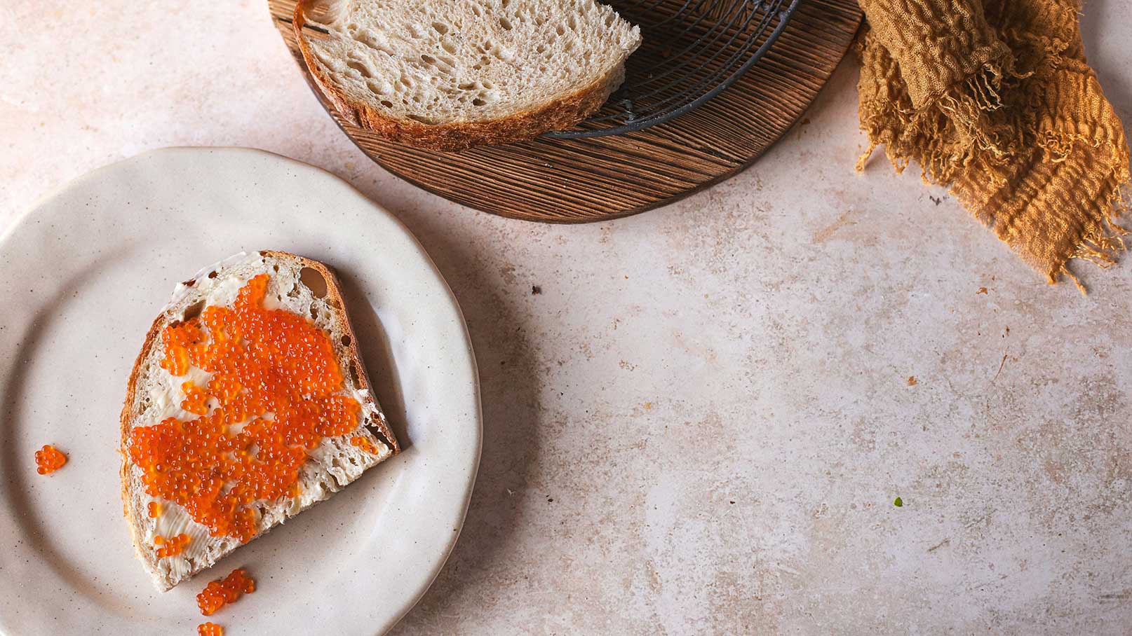 Ein Stück Brot mit einem Aufstrich aus Frischkäse, das mit orangefarbenen Kaviarperlen dekoriert ist. Der Teller ist hell und rund, die Umgebung zeigt einen rustikalen Stil mit einem weiteren Brotschnitt und einem Textilstück in der Ecke.