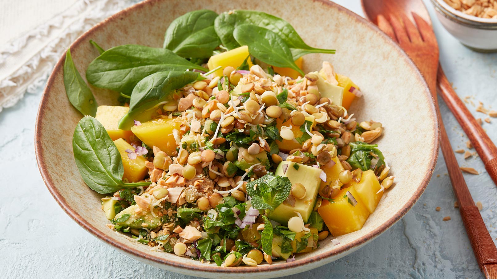 Eine Schüssel mit buntem Salat, der aus frischem Spinat, gelben Mangostücken, Linsen, Kräutern und gerösteten Erdnüssen besteht. Die Zutaten sind gut vermischt und verleihen dem Gericht eine abwechslungsreiche Textur und einen ansprechenden Geschmack.