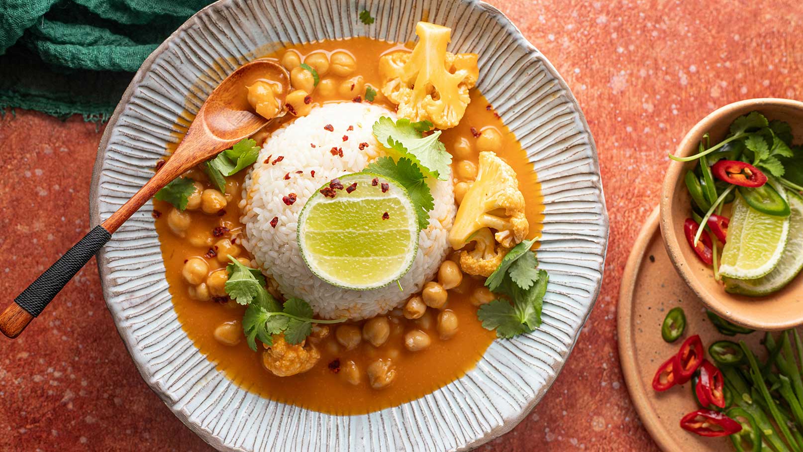 Ein Teller mit Basmati-Reis, umgeben von Kichererbsen und Blumenkohl in einer orangefarbenen Sauce. Frische Kräuter und eine Limettenscheibe dekorieren das Gericht, das mit roten Chiliflocken bestreut ist. Der Teller hat eine leicht gewellte Form und steht auf einem rustikalen Untergrund.