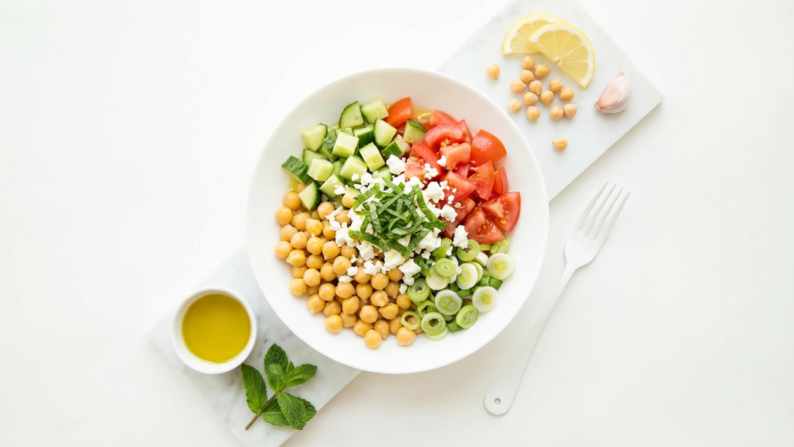 Eine Schüssel mit Kichererbsen, gewürfeltem Gurken, Tomaten, Frischkäse und gehacktem Minze. Um die Schüssel herum liegen Olivenöl, frische Minze, Zitronenscheiben, und eine Knoblauchzehe auf einem weißen Untergrund.