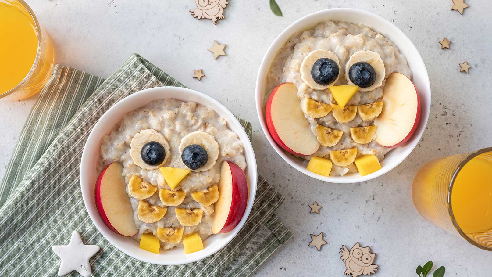 Zwei Schalen Haferbrei sind kreativ zu Eulen gestaltet. Bananen, Äpfel und Heidelbeeren bilden die Augen, den Schnabel und die Ohren. Neben den Schalen steht ein Glas Orangensaft. Der Hintergrund ist hell mit kleinen Deko-Elementen.