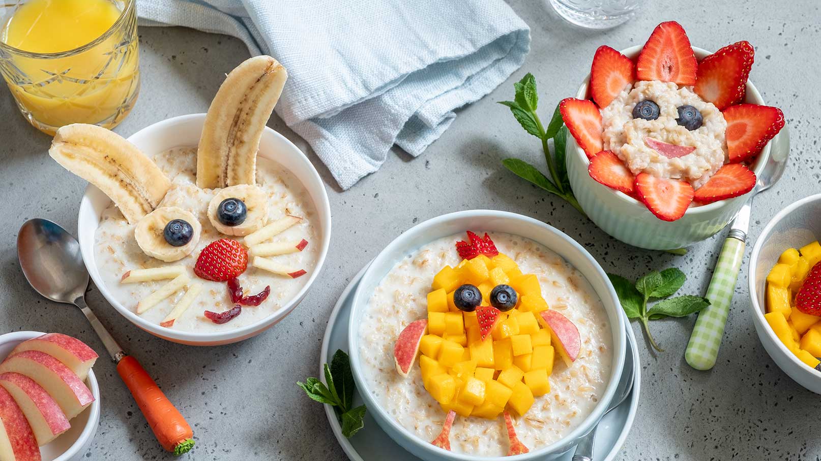 Zwei Schalen mit dekoriertem Haferbrei, eine in Form eines Hasen mit Bananenohren und eine andere als Küken mit Mangostücken. Eine dritte Schale zeigt einen blumenähnlichen Haferbrei, geschmückt mit Erdbeeren und Heidelbeeren. Daneben liegen geschnittene Äpfel und eine Karotte.