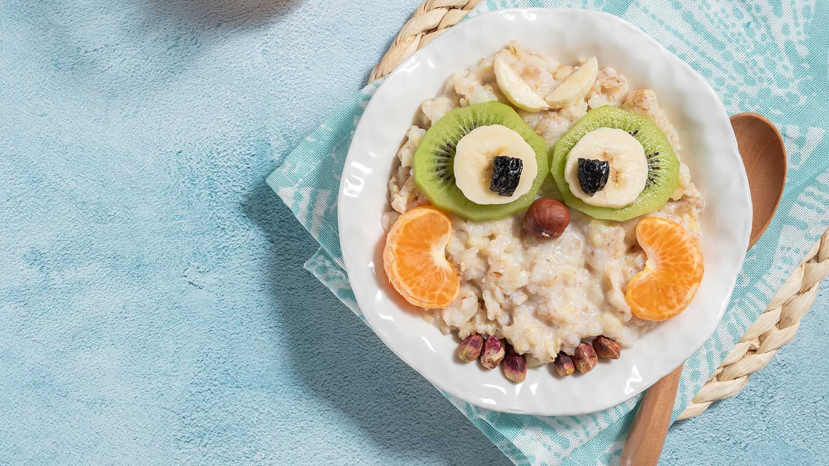 Eine Schüssel Haferbrei dekoriert mit Fruchtstücken, die wie ein Gesicht aussehen. Kiwi- und Bananenscheiben bilden die Augen, ergänzt durch eine Getrocknete Frucht als Pupille. Mandarinen- und Haselnussstücke verweisen auf einen freundlichen Ausdruck. Serviert auf einem blauen Untergrund mit einem Löffel daneben.