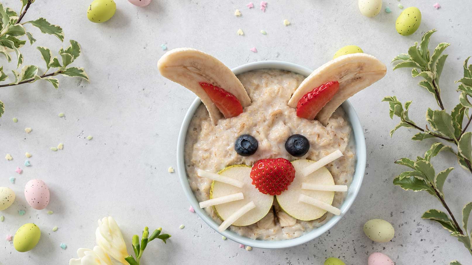 Eine Schüssel Haferbrei, dekoriert als Hase. Die Ohren bestehen aus Bananenscheiben, mit Erdbeeren. Blaubeeren dienen als Augen und eine Erdbeere als Nase. Apfelscheiben stellen den Mund und die Schnurrhaare dar. Bunte Schokoladeneier und Pflanzenranken umrahmen die Schüssel.