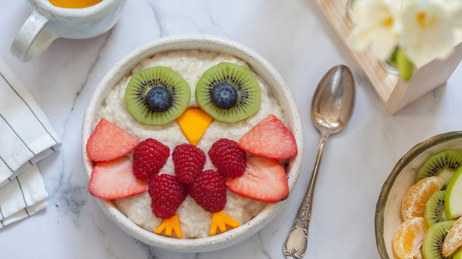 Eine Schüssel mit Haferbrei, der kreativ wie eine Eule dekoriert ist. Die Augen bestehen aus Kiwi und Blaubeeren, der Schnabel aus Mango. Himbeeren und Erdbeeren formen den Körper der Eule. Im Hintergrund sind ein Löffel und eine Tasse Tee zu sehen.