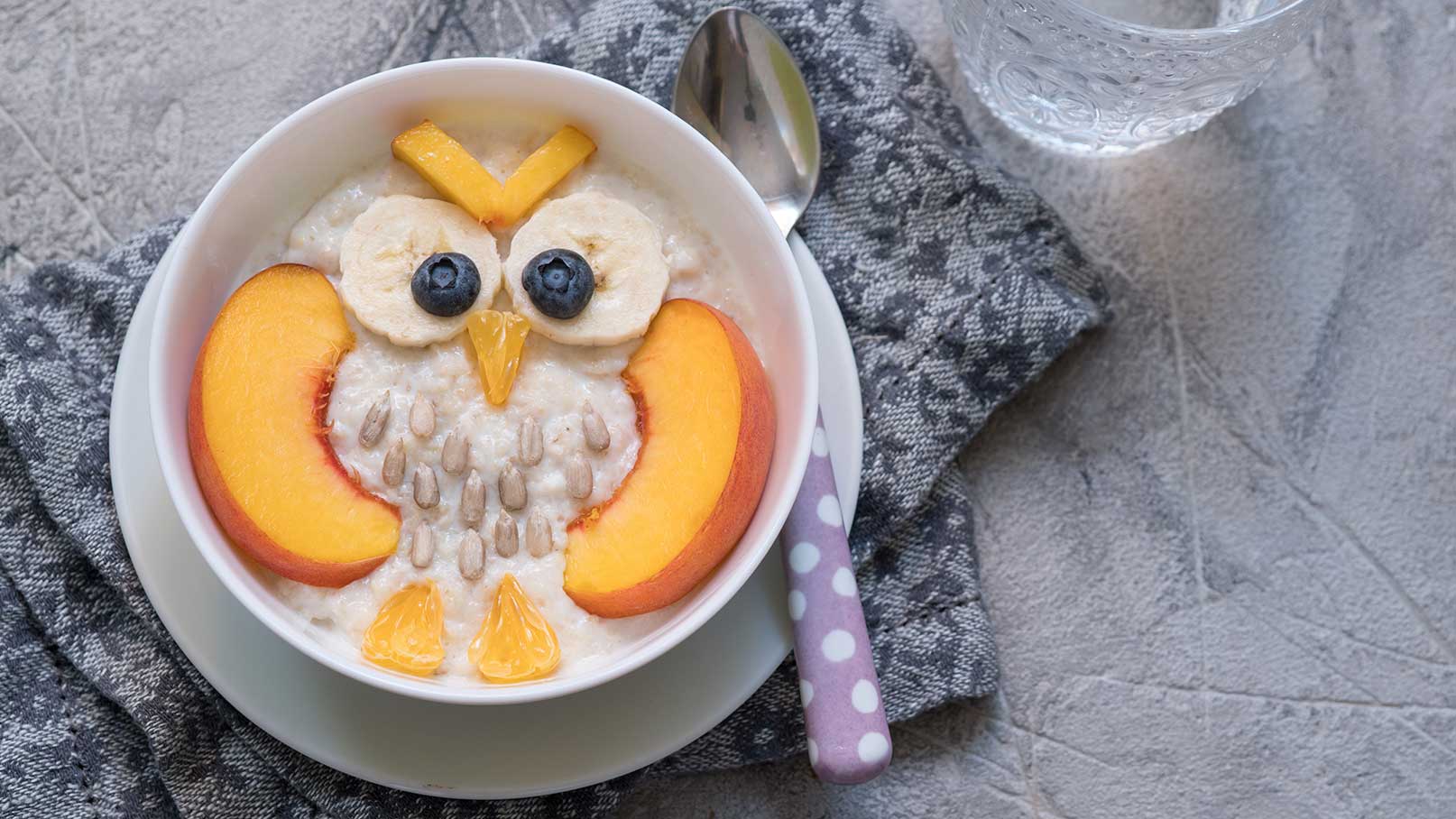Eine Schüssel Haferbrei ist kreativ angerichtet und sieht aus wie eine Eule. Die Augen sind aus Bananenscheiben und Heidelbeeren, mit einer Nase aus einer kleinen Karotte. Die Flügel sind aus Pfirsichscheiben, während Sonnenblumenkerne den Körper bilden. Die Schüssel steht auf einem grauen Tuch.