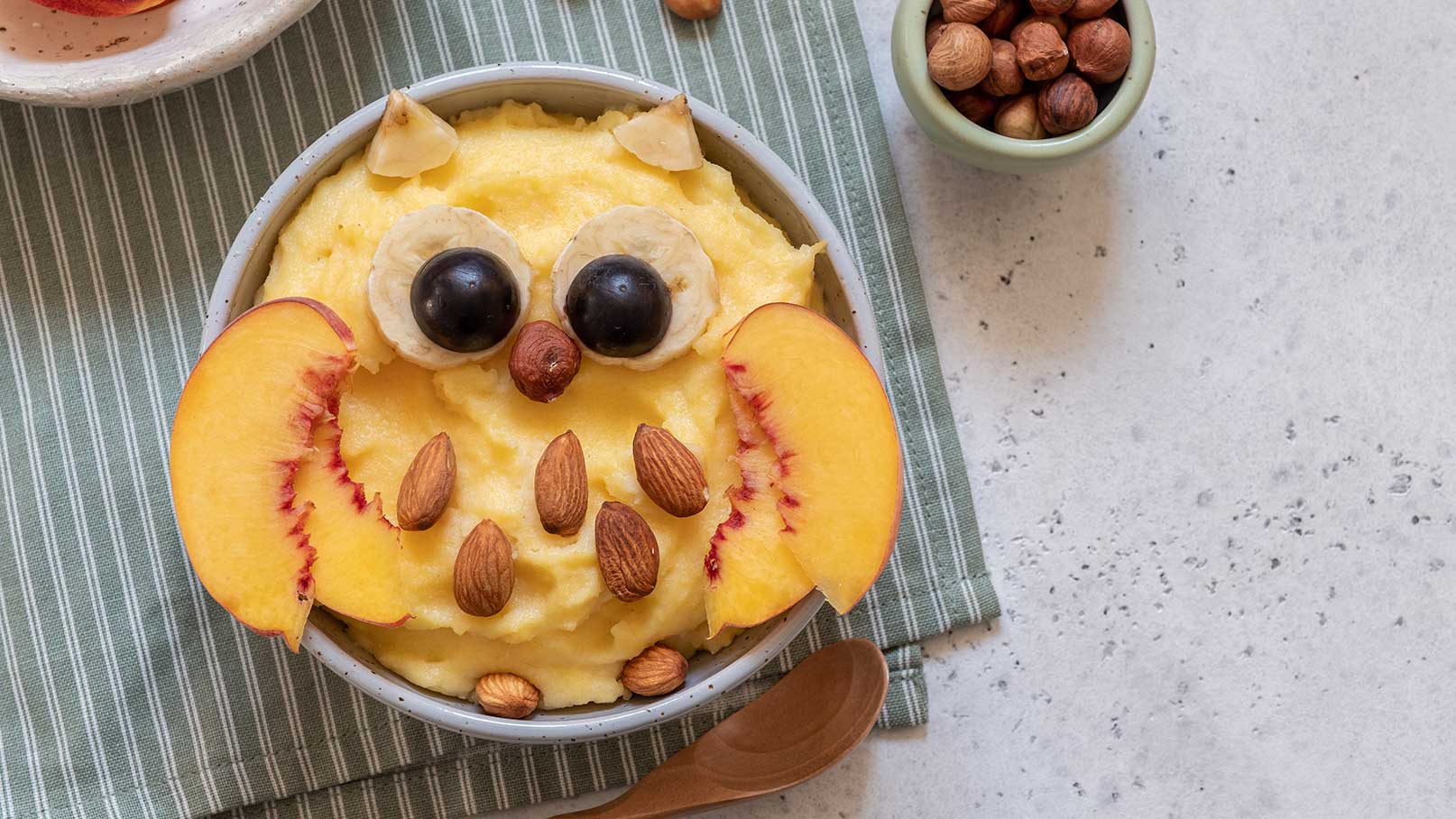 Eine Schüssel mit Püree, dekoriert als Eule. Die Eule hat Ohren aus Pfirsichscheiben, Augen aus Bananen und Heidelbeeren sowie eine Nase aus einer Nuss. Mandeln sind als Federn platziert. Im Hintergrund sind eine grüne Tischdecke und eine kleine Schüssel mit Nüssen.